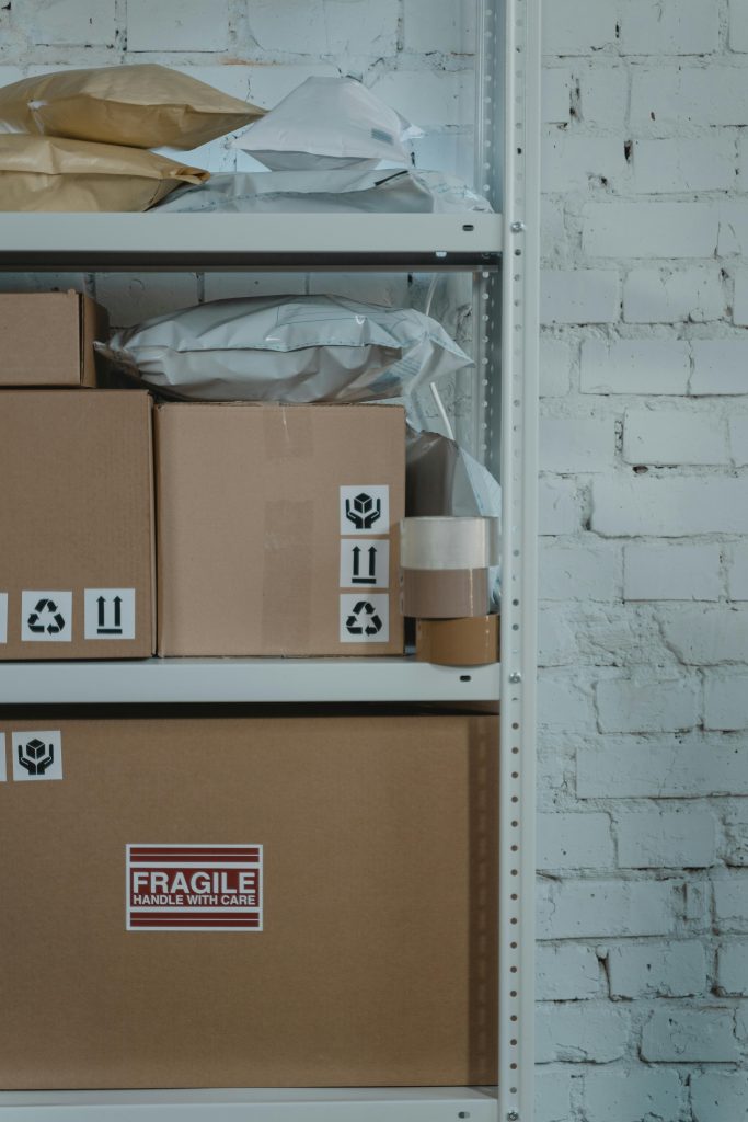 A warehouse shelf with labeled cardboard boxes and packing materials against a brick wall.
